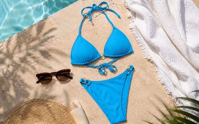 Blue bikini flat lay by a swimming pool with sunglasses, sunscreen, a white towel, and a straw hat on a sunny pool deck.