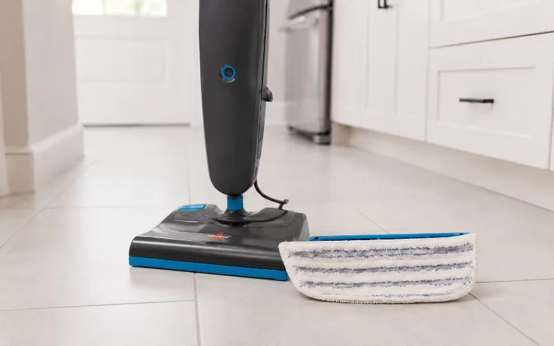 BISSELL Steam & Sweep mop pad with blue stripes resting beside the machine base on light tile flooring.