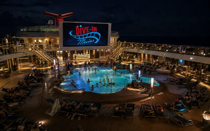 Passengers watching an outdoor movie from lounge chairs surrounding a cruise ship pool at night.