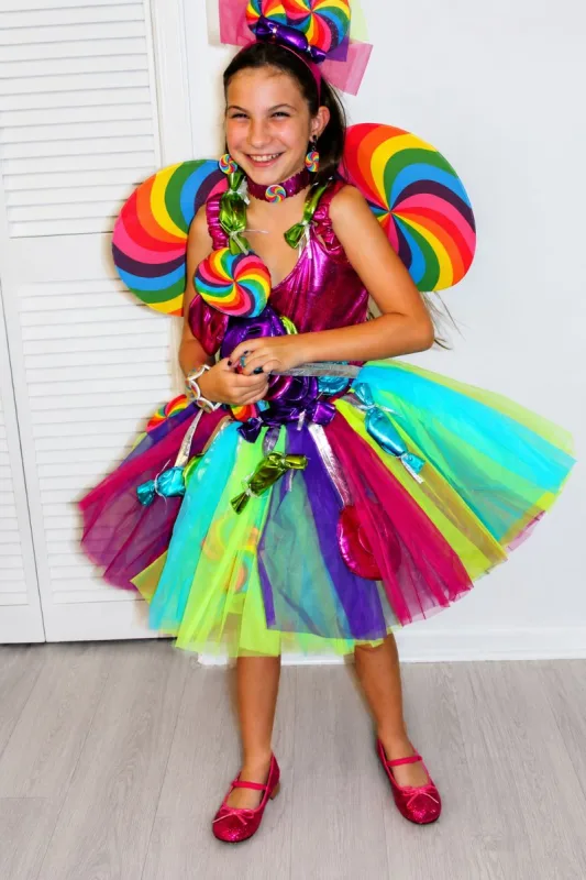 Girl wearing a colorful Candy Fairy costume with rainbow lollipop wings, a bright layered tulle skirt, candy jewelry, and pink shoes.