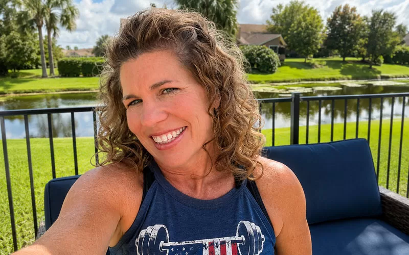 Woman with curly hair smiling outdoors by a lake wearing a navy graphic tank top in a bright sunny backyard setting.