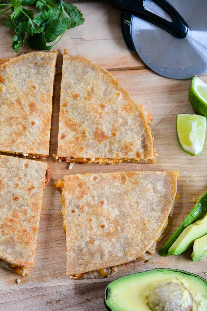 Sliced rotisserie chicken quesadillas on a wooden cutting board with avocado, lime wedges, cilantro, and a pizza cutter.