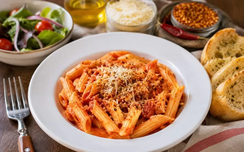 Spicy marinara penne pasta served with garlic bread, green salad, Parmesan cheese, and red pepper flakes.