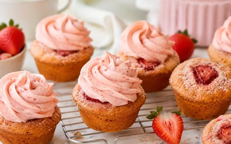 Strawberry cupcakes with pink strawberry frosting, baked strawberry slices, and fresh strawberries on a cooling rack