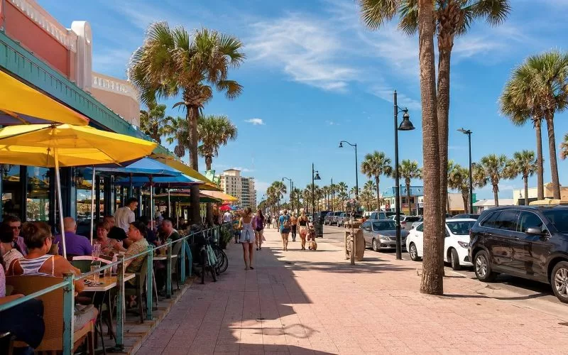 Daytona Beach street scene with outdoor dining, colorful umbrellas, palm trees, people walking, and a sunny coastal vacation feel.