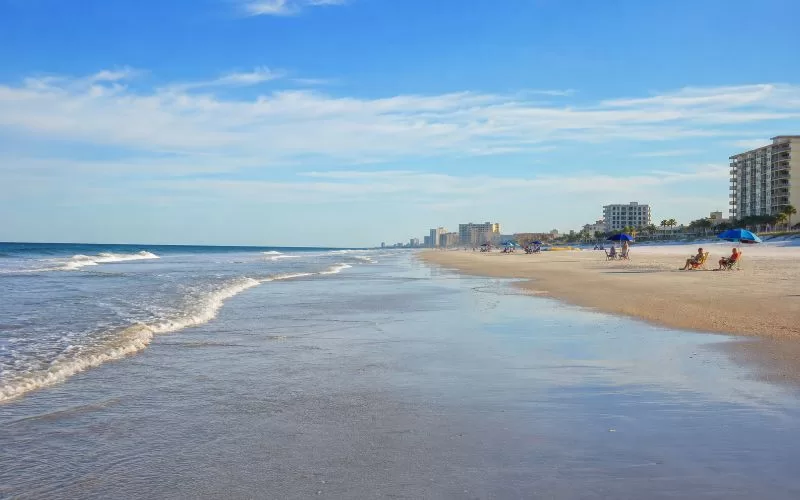 Peaceful Daytona Beach shoreline with soft waves, open wet sand, distant umbrellas, and beachfront buildings in warm daylight.