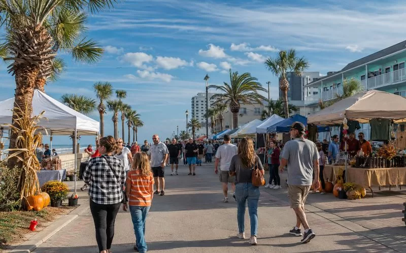 Outdoor fall festival near the Daytona Beach coast with vendor tents, palm trees, pumpkins, and people walking along the promenade.