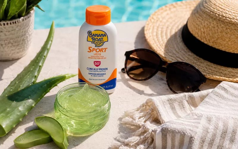 Aloe vera gel, aloe leaves, Banana Boat sunscreen, sunglasses, straw hat, and towel arranged beside a swimming pool.