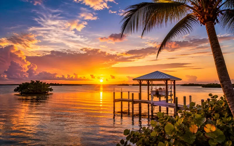 Golden sunset over calm Marco Island water with a dock, palm tree, and colorful clouds