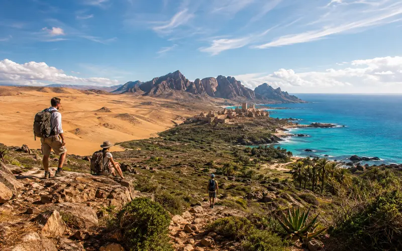 Travelers exploring a scenic African-inspired landscape with desert terrain, mountains, greenery, and coastline.