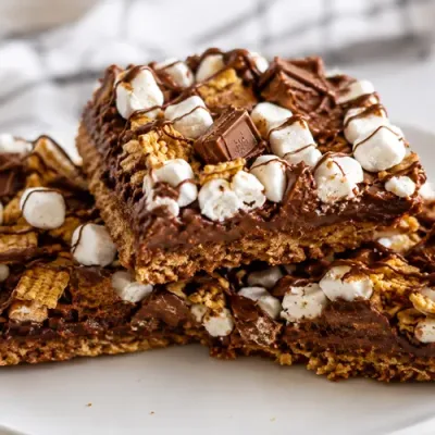 Golden Grahams S’mores Pizza Squares stacked on a white plate with marshmallows, chocolate pieces, cereal, and chocolate drizzle.