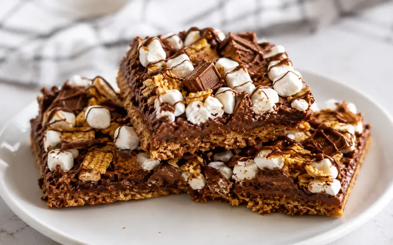 Golden Grahams S’mores Pizza Squares stacked on a white plate with marshmallows, chocolate pieces, cereal, and chocolate drizzle.