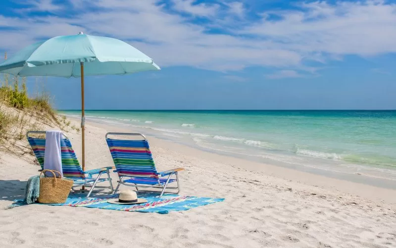 Two beach chairs, an umbrella, towels, and a beach bag set up on a quiet Gulf Coast beach.
