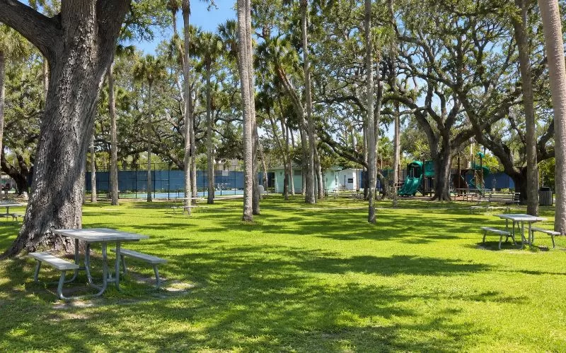 Shaded Florida park with picnic tables, palm trees, tennis courts, playground equipment, and green grass.