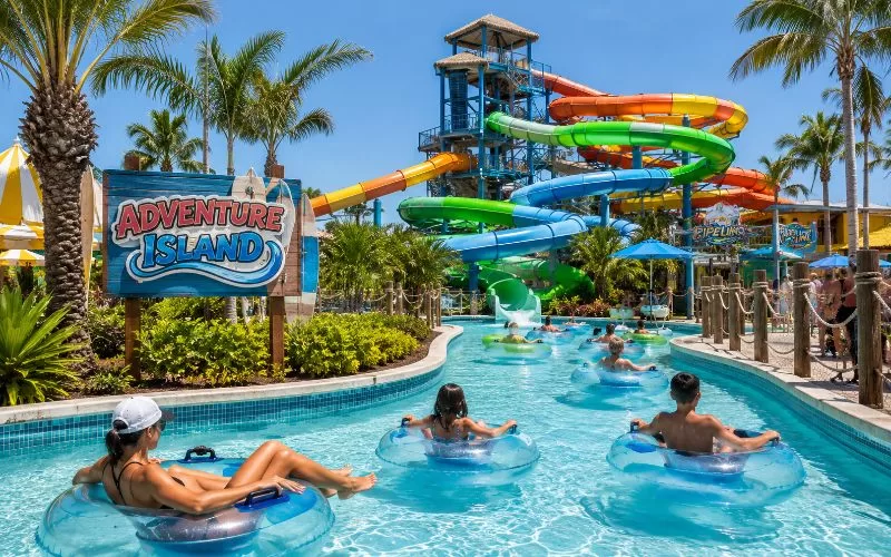 Families floating on tubes through a sunny Tampa water park lazy river with colorful slides and palm trees in the background.