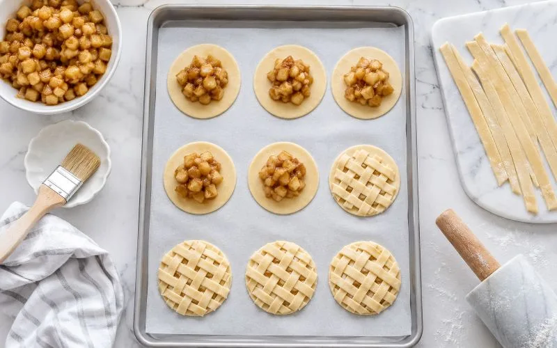 Caramel apple pie cookies being assembled with fresh cinnamon apple filling, round pie crust bases, lattice tops, and strips of pie dough on a baking sheet.