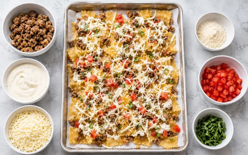 Lasagna noodle pieces being assembled with Italian sausage, Alfredo sauce, mozzarella, Parmesan, tomatoes, and basil on a baking sheet.