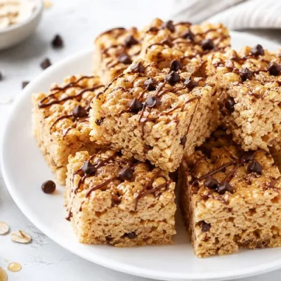 Healthy rice krispie treats with peanut butter, mini chocolate chips, and a light chocolate drizzle stacked on a white plate with oats, honey, and peanut butter nearby.