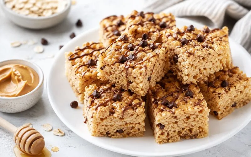 Healthy rice krispie treats with peanut butter, mini chocolate chips, and a light chocolate drizzle stacked on a white plate with oats, honey, and peanut butter nearby.