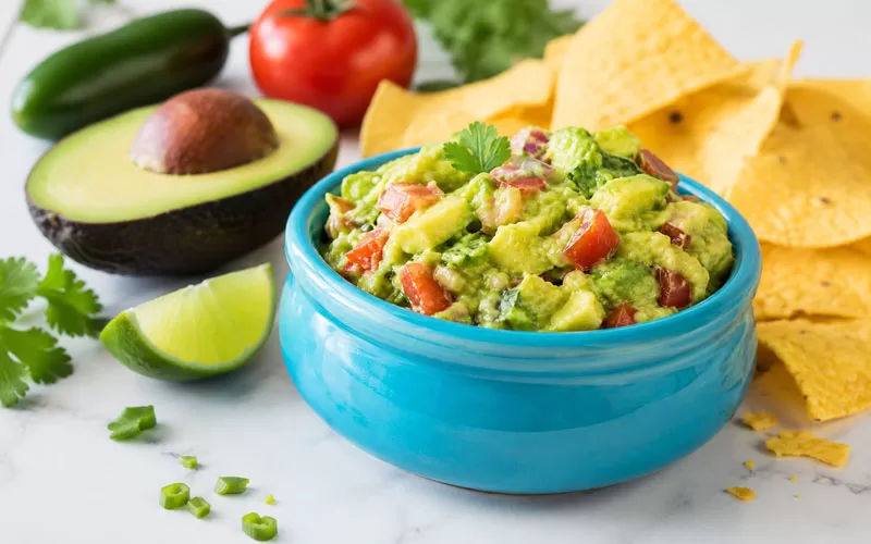 Turquoise bowl of chunky avocado salsamole guacamole dip with tortilla chips, lime, avocado, tomato, and jalapeño on a light countertop.