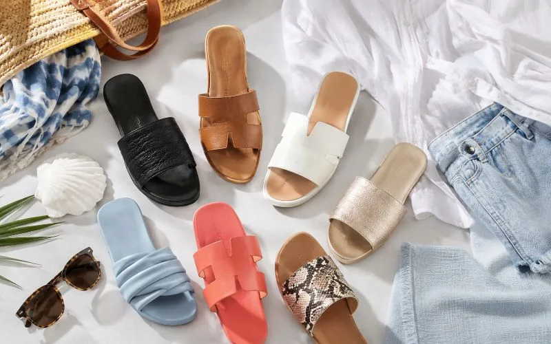 Flat lay of women’s slide sandals in black, tan, white, metallic gold, soft blue, coral, and patterned styles with summer clothing and accessories.