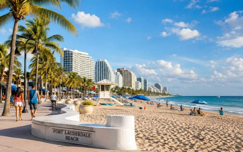 Fort Lauderdale beach promenade with palm trees, beachfront skyline, lifeguard tower, and people walking along the oceanfront.