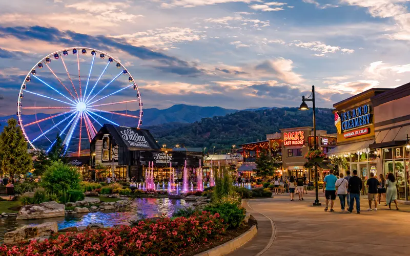 Great Smoky Mountain Wheel illuminated at The Island in Pigeon Forge with fountains, shops, visitors, and Smoky Mountains at dusk.