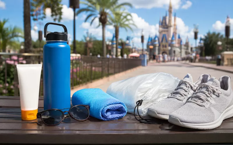 Theme park day essentials with sunscreen, sunglasses, cooling towel, refillable water bottle, poncho, and walking shoes on a sunny bench.