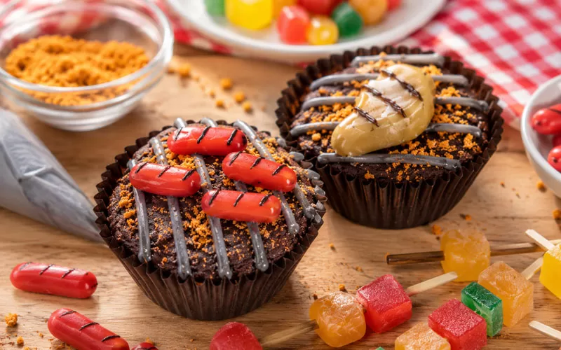 Close-up of two Father’s Day Grillin’ Brownies decorated like mini grills, with candy hot dogs, a candy steak, frosting grill lines, and extra decorating pieces scattered on the table.