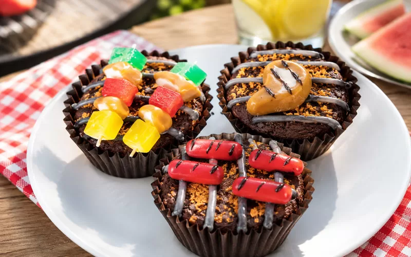 Plate of Father’s Day Grillin’ Brownies decorated like mini barbecue grills with candy kabobs, hot dogs, and a steak on a picnic table outdoors.