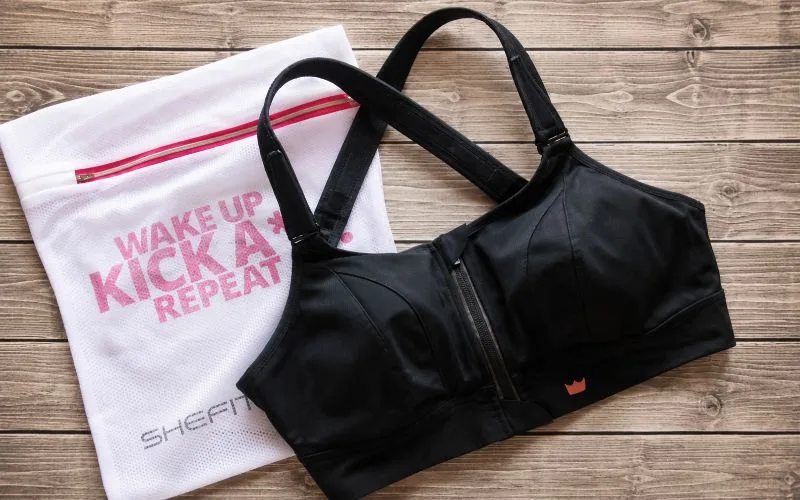 Black SHEFIT Ultimate Sports Bra displayed beside a white mesh laundry bag on a wood surface.