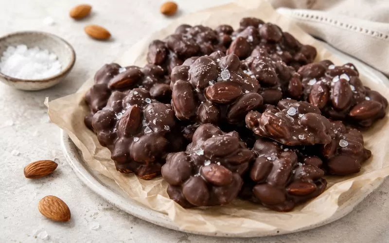 Dark chocolate almond clusters sprinkled with flaky sea salt on parchment with whole almonds and a small bowl of salt nearby.