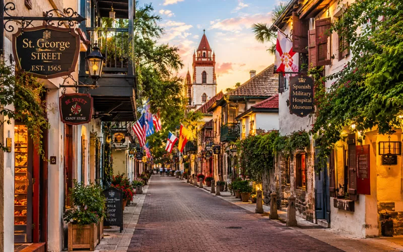 Historic downtown St. Augustine street with cobblestone road, Spanish-style buildings, shop signs, flags, and warm golden evening light.