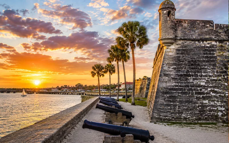 Castillo de San Marcos at sunset with cannons, palm trees, waterfront views, and glowing skies over St. Augustine.