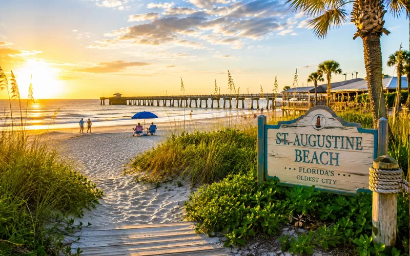 St. Augustine Beach at sunset with colorful beach chairs, umbrellas, palm trees, boardwalk path, and a scenic pier in the background.