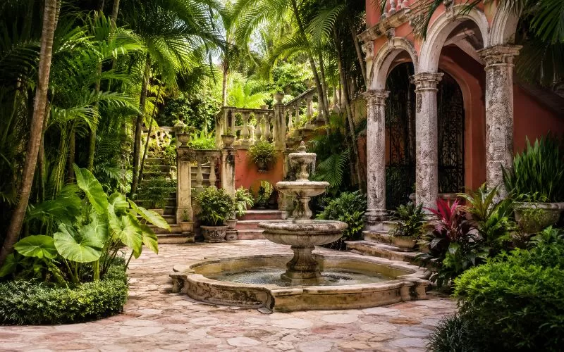 Elegant tropical garden courtyard with stone pathways, lush greenery, architectural arches, and a fountain.