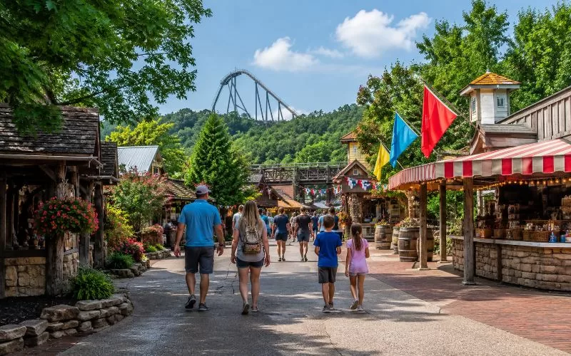 Family walking through a rustic Ozarks-inspired attraction area with colorful flags, greenery, and a ride in the distance.