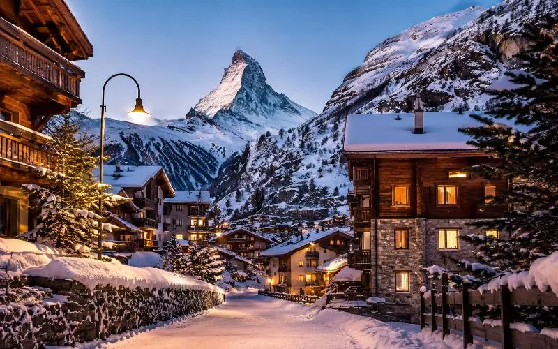 Snowy Swiss alpine village at twilight with chalet lights, mountain peaks, and a Matterhorn-inspired peak in the distance.