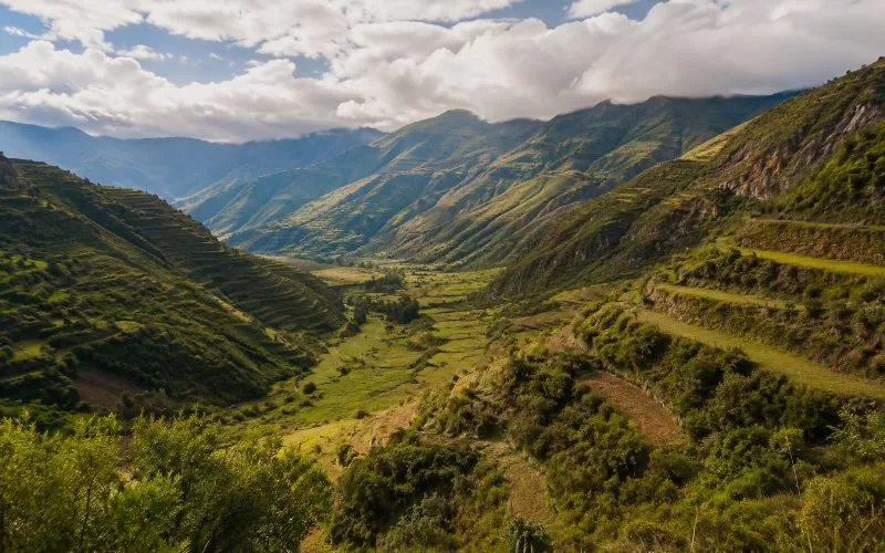 Scenic Andean valley landscape with green mountain slopes, terraced hillsides, winding paths, and soft clouds.