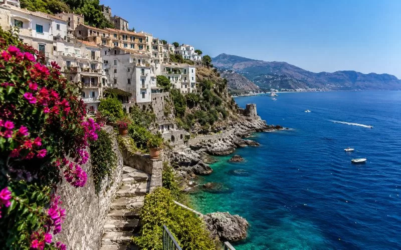 Cliffside coastal village with white buildings, stone steps, pink flowers, and bright blue Mediterranean water.