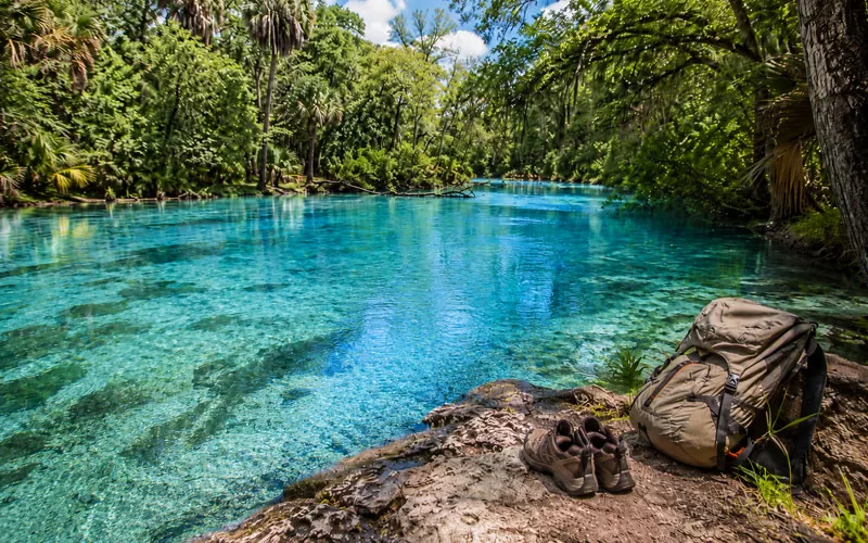 Clear blue Florida spring water surrounded by trees with hiking shoes and a backpack on the riverbank.