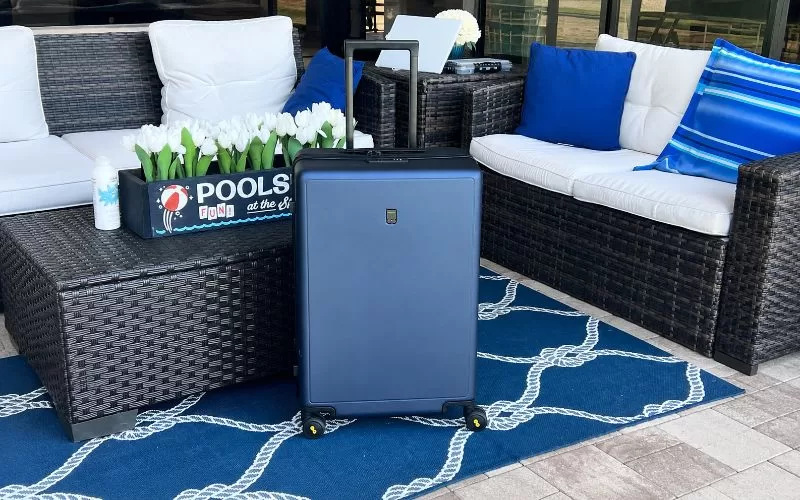 Navy LEVEL8 suitcase on a blue outdoor rug surrounded by white patio cushions, blue pillows, wicker furniture, and white flowers.