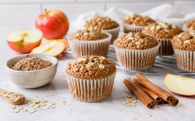 Freshly baked apple cinnamon flaxseed muffins with apples, oats, cinnamon sticks, and ground flaxseed on a light kitchen counter.