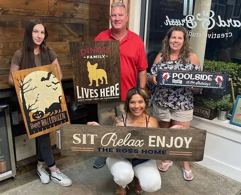 Family members holding finished Board & Brush wood craft projects inside the studio.
