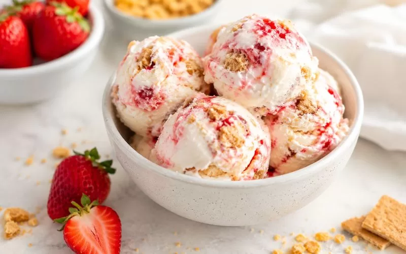 Bowl of homemade strawberry cheesecake ice cream with strawberry swirls, graham cracker pieces, fresh strawberries, and graham cracker crumbs on a light counter.