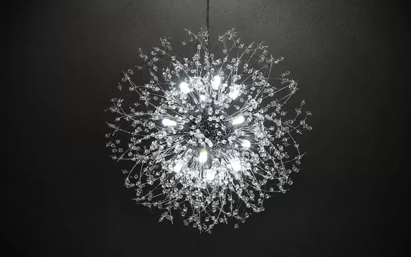 Lit crystal dandelion chandelier against a dark ceiling, showing the exposed bulbs, chrome arms, and crystal bead details.