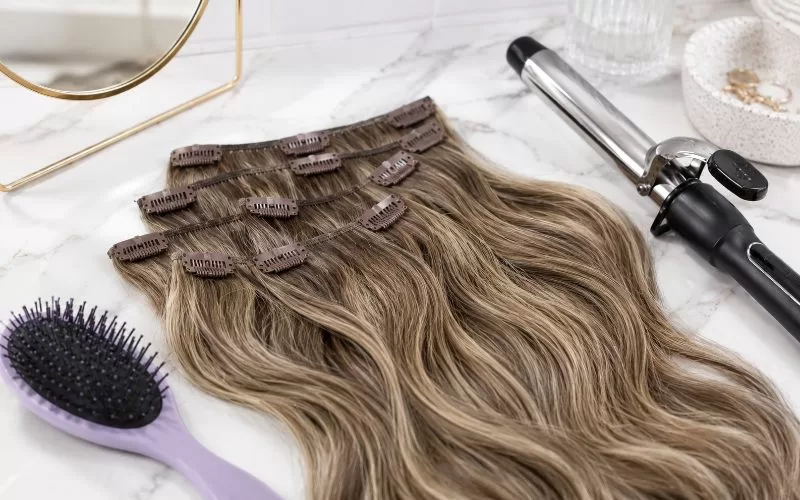 Clip-in caramel balayage hair extensions on a white marble vanity with a purple hairbrush and curling iron.