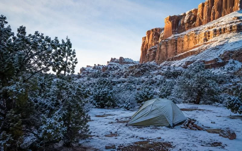 Tent campsite below snow-dusted red rock cliffs with evergreen shrubs, pale winter sunlight, and cool blue-gray shadows.