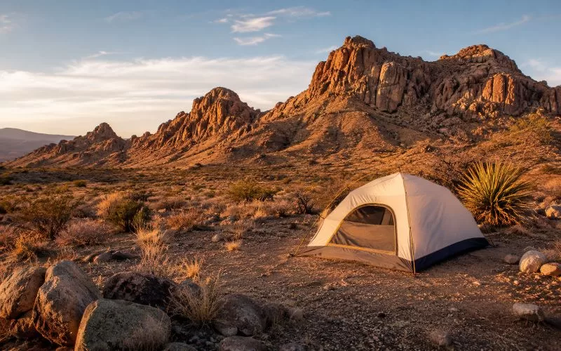 Tent campsite in a rugged desert landscape with dry grasses, rocky mountains, and warm golden-hour light.