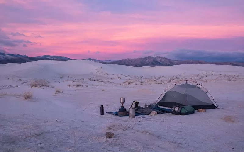 Tent campsite near pale desert dunes and distant rocky mountains under a lavender-pink evening sky.